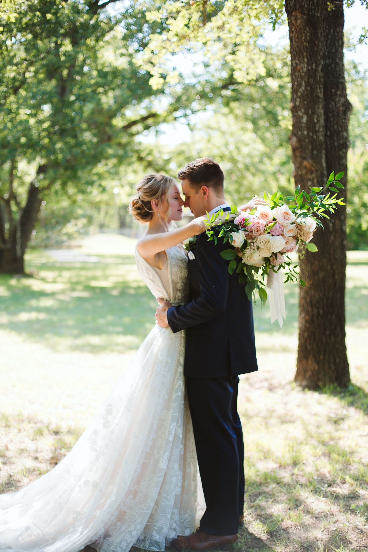 Newlyweds embrace under tree canopy in sunlit outdoor wedding portrait.