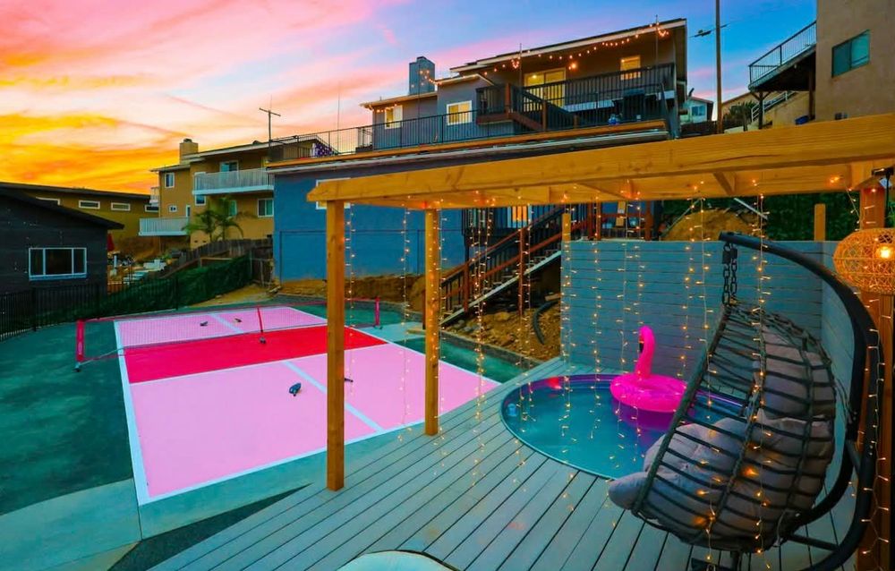Modern rooftop deck with curved wooden flooring, pink accent wall, and string lights under a vibrant sunset sky.