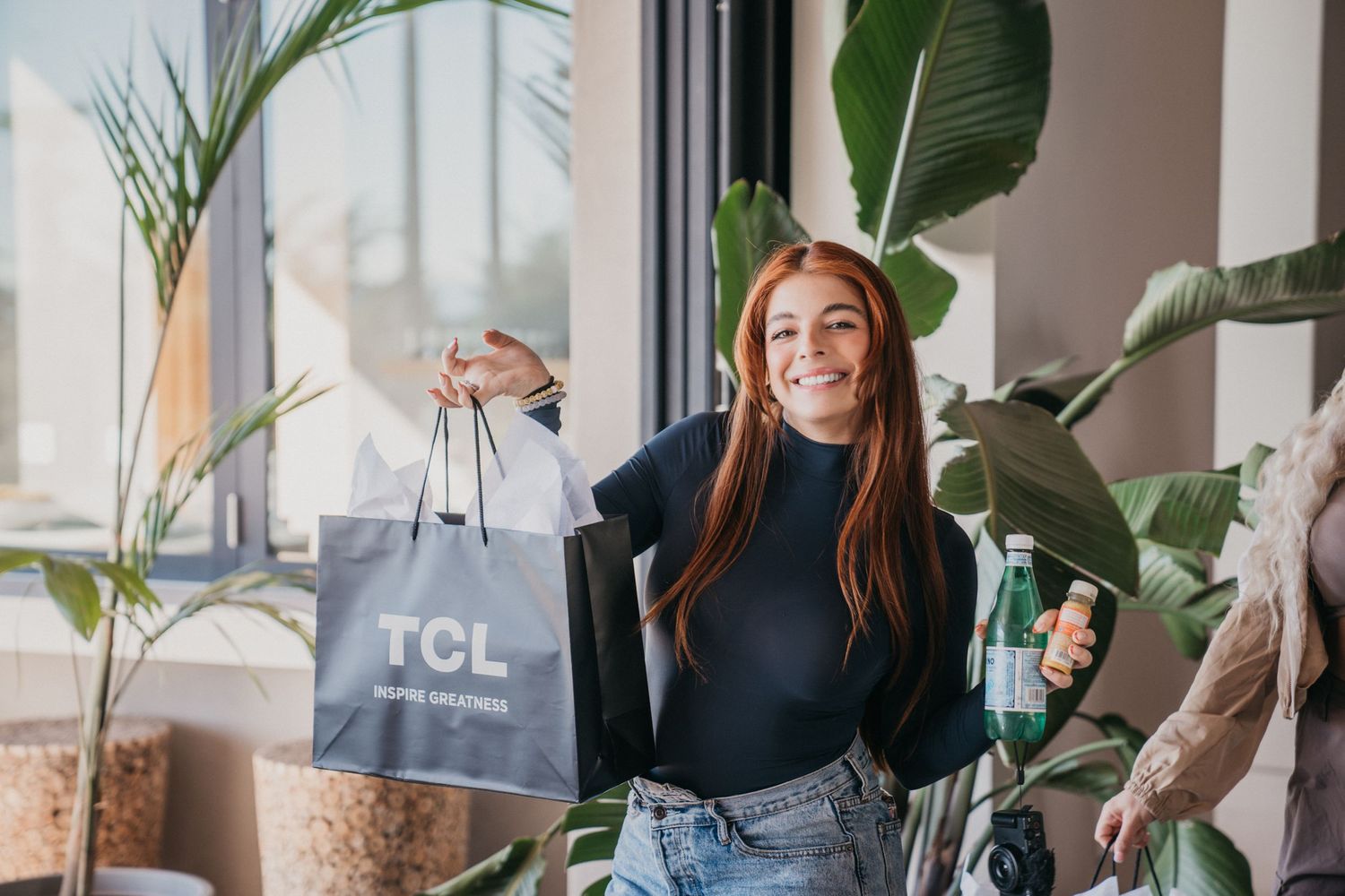 Woman holding shopping bag while posing with indoor plants in bright retail space.