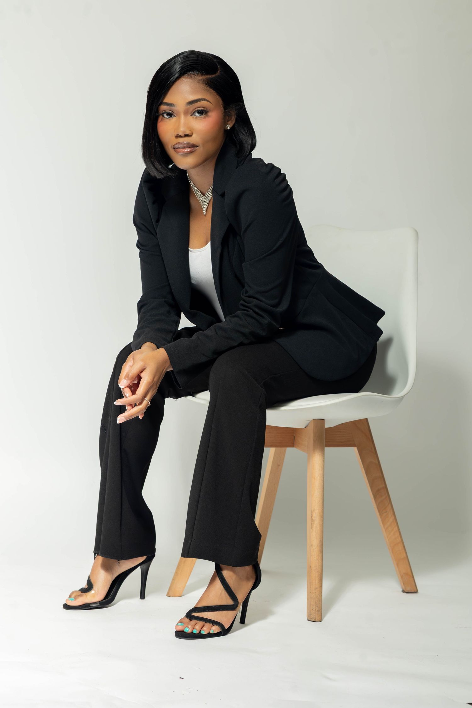 Young woman in black blazer and heels seated on white chair against light background.