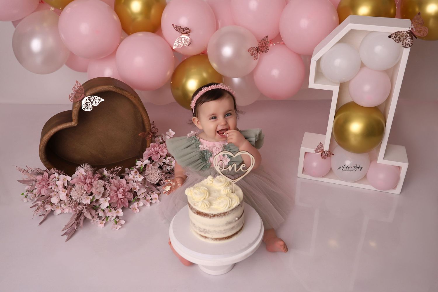 First birthday cake smash setup with pink and gold balloons, heart decor, and white cake on pink background.