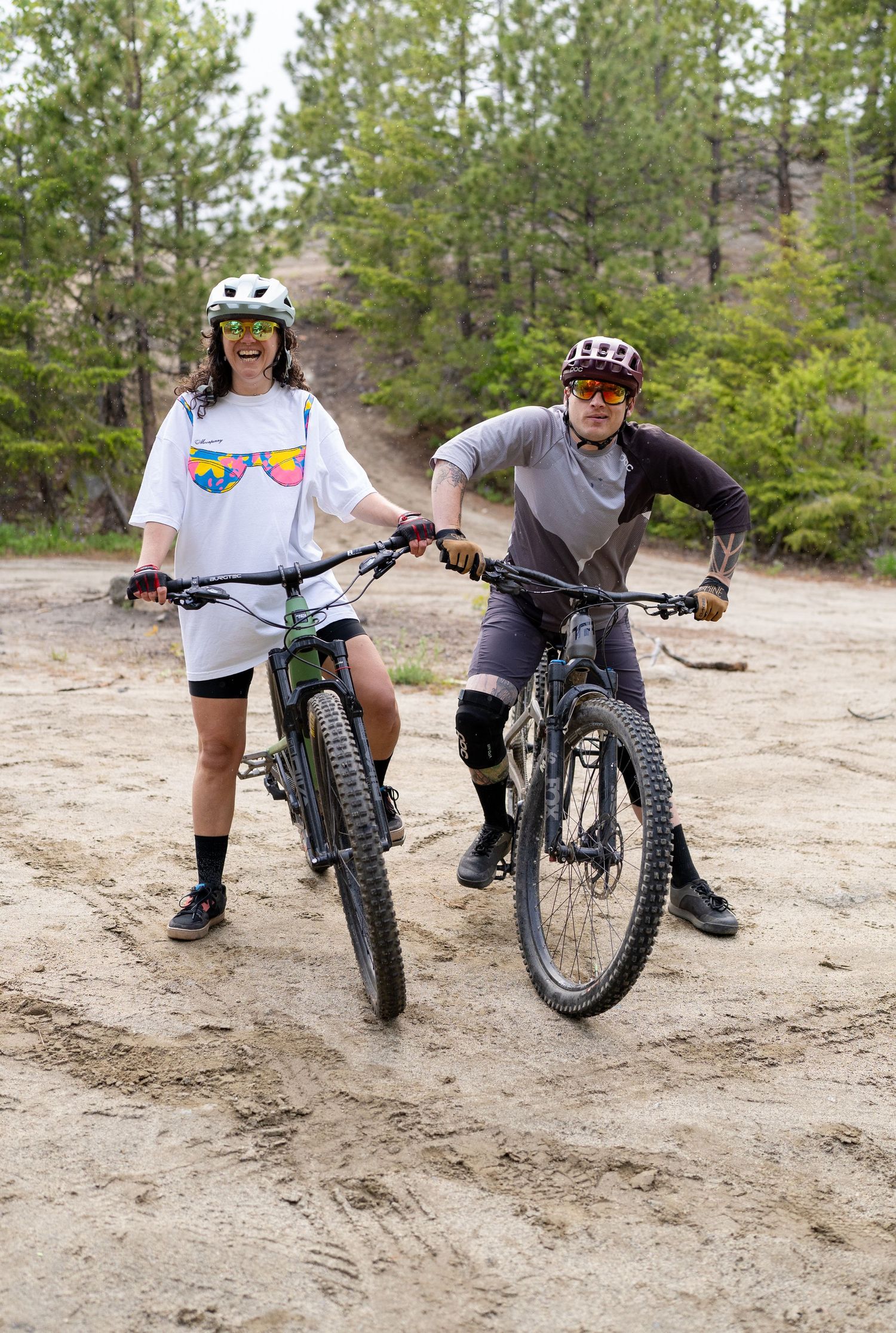 Two mountain bikers with helmets pose with their bikes on a dirt trail.
