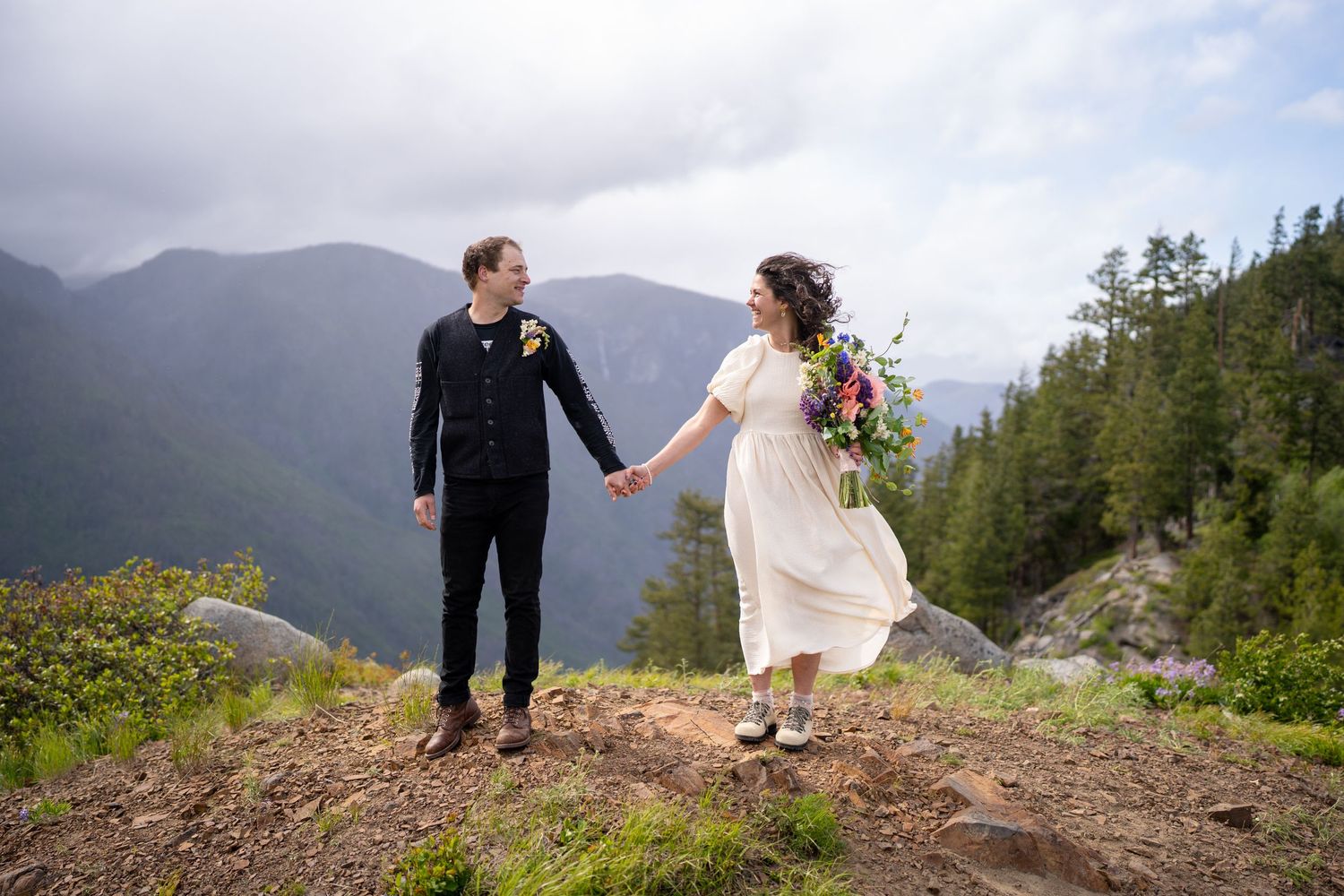 Couple holds hands on mountain peak with dramatic landscape views.