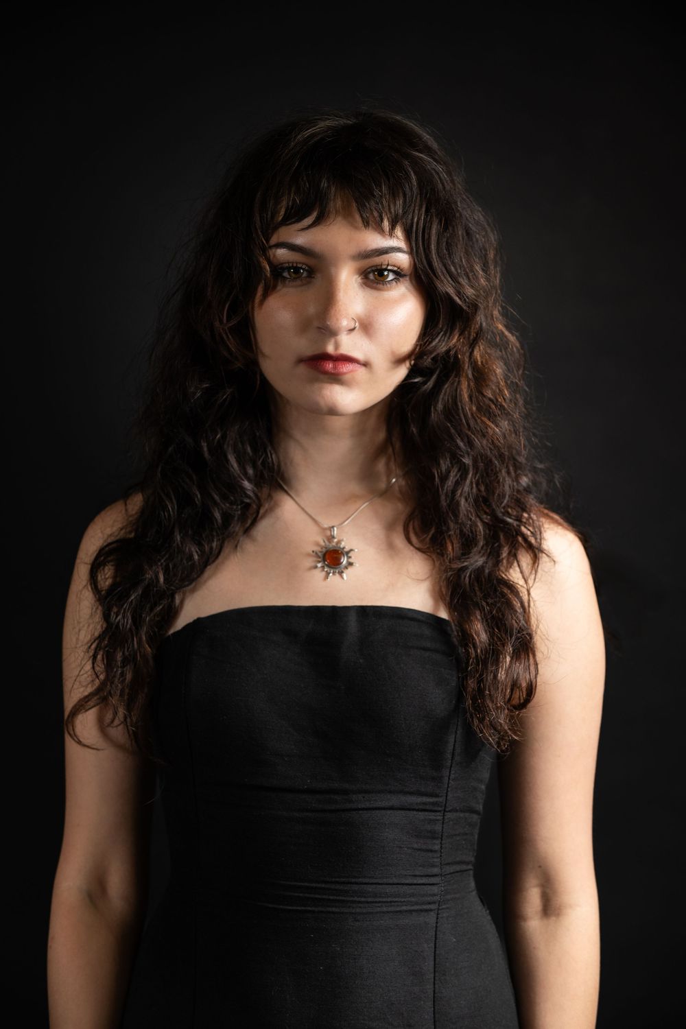 Portrait against dark background shows model in black sleeveless dress with curly dark hair.