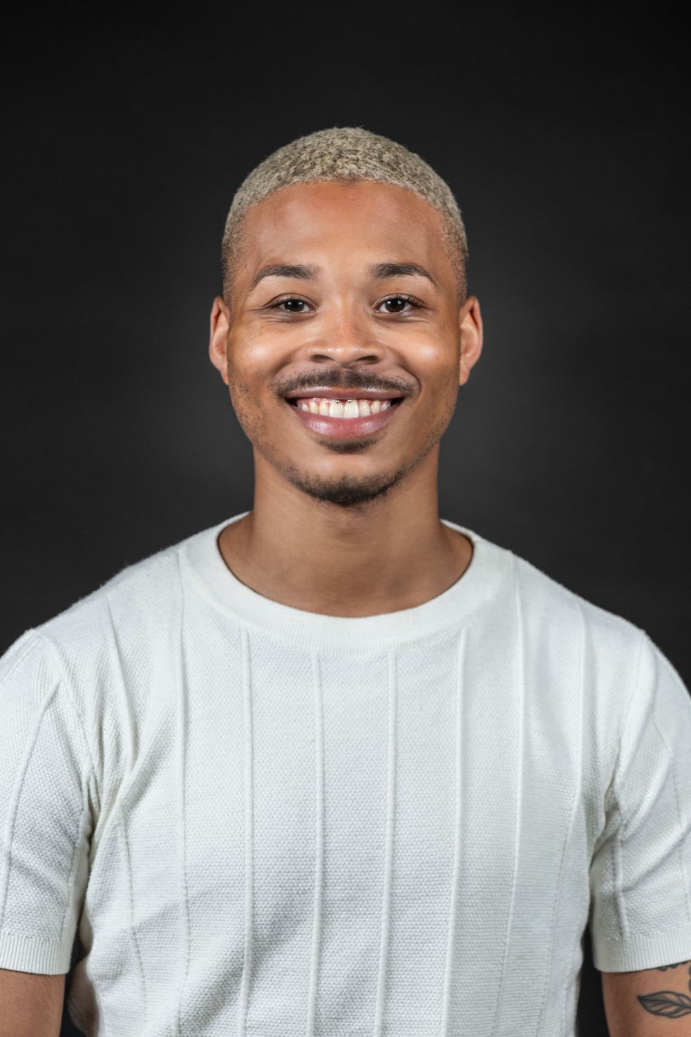 Portrait of a smiling person with bleached blonde hair and arm tattoo wearing a white t-shirt against dark background.