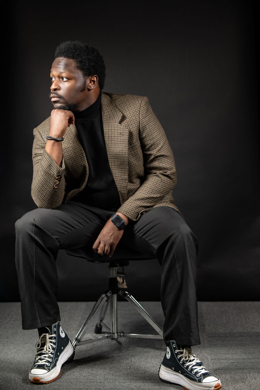 Professional portrait in a plaid blazer and black turtleneck sitting thoughtfully on a stool against dark background.