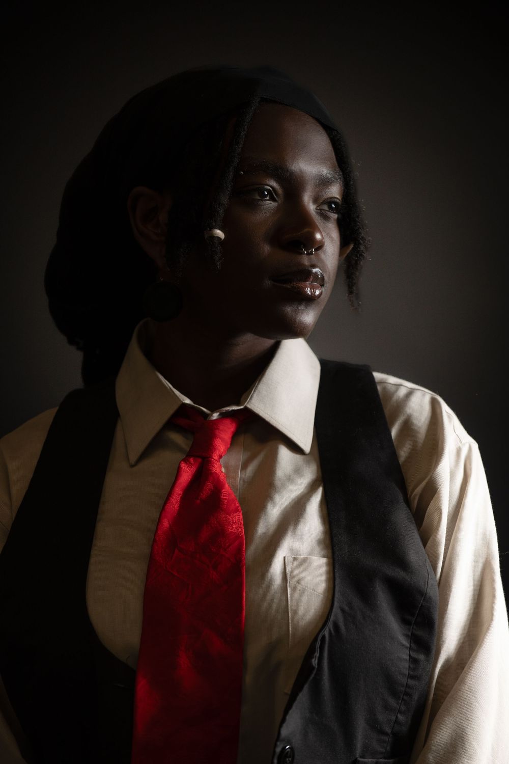 A series of dramatic portraits shot in moody lighting featuring a person wearing a black vest, white shirt and bright red tie.