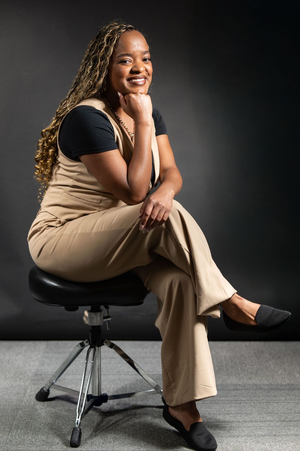Portrait of someone sitting on black office chair wearing black top and beige overalls against dark background.