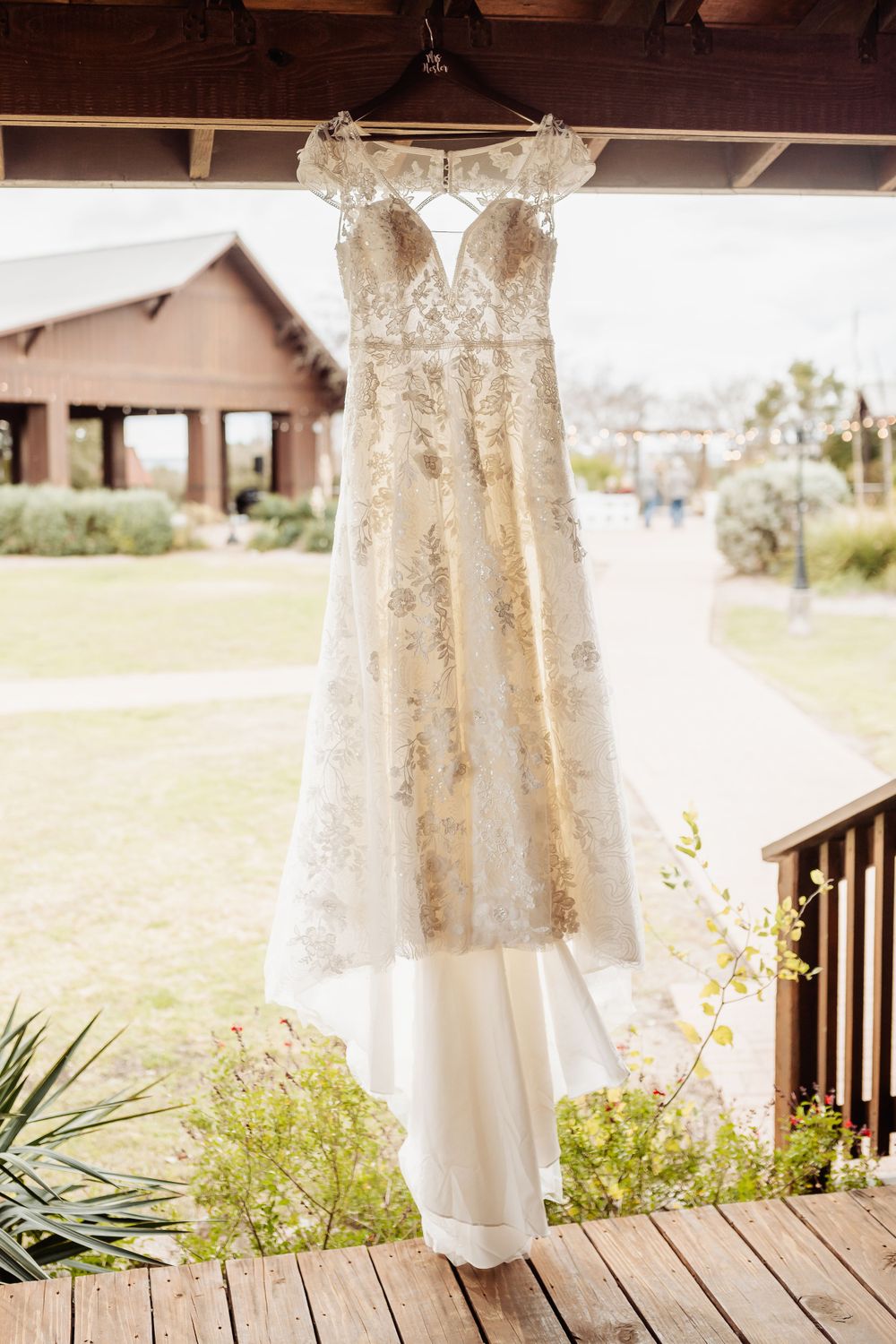Lace wedding dress hanging on wooden porch with rustic venue in background.
