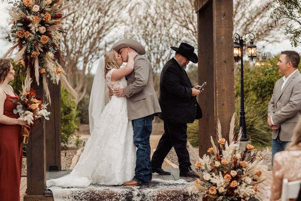 Couple embracing during rustic outdoor wedding ceremony under wooden arch with peach florals.