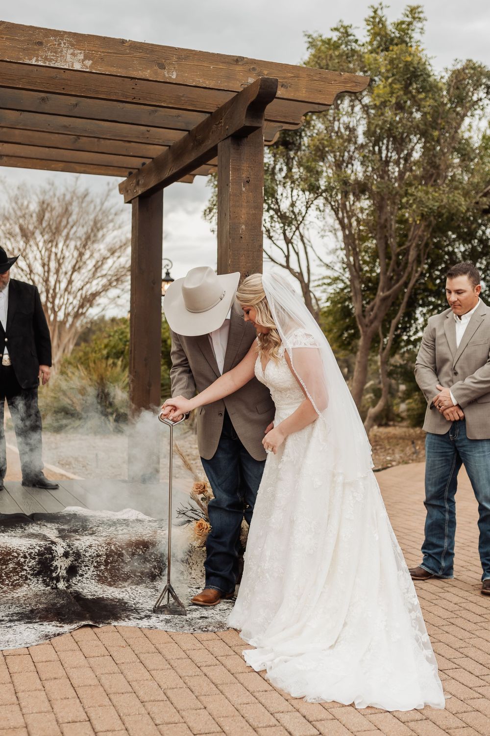 Couple performing a wedding ritual under a wooden pergola with smoke rising from the ground during an outdoor ceremony.