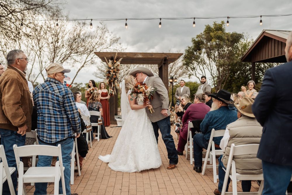 Wedding ceremony taking place under a rustic wooden pergola with string lights and white chairs.