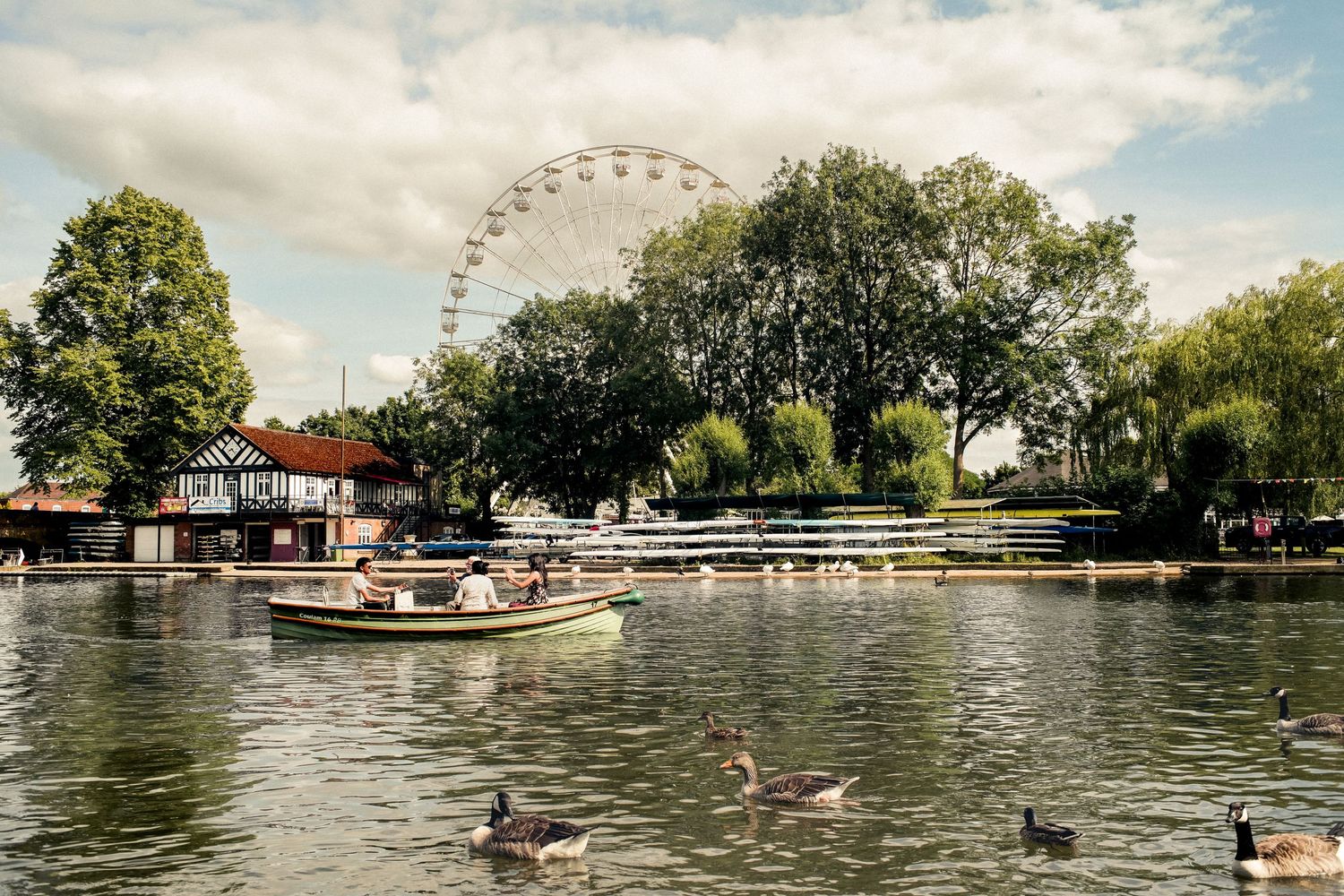 A scenic lake view with boats, ducks, and a ferris wheel on the horizon under cloudy skies.