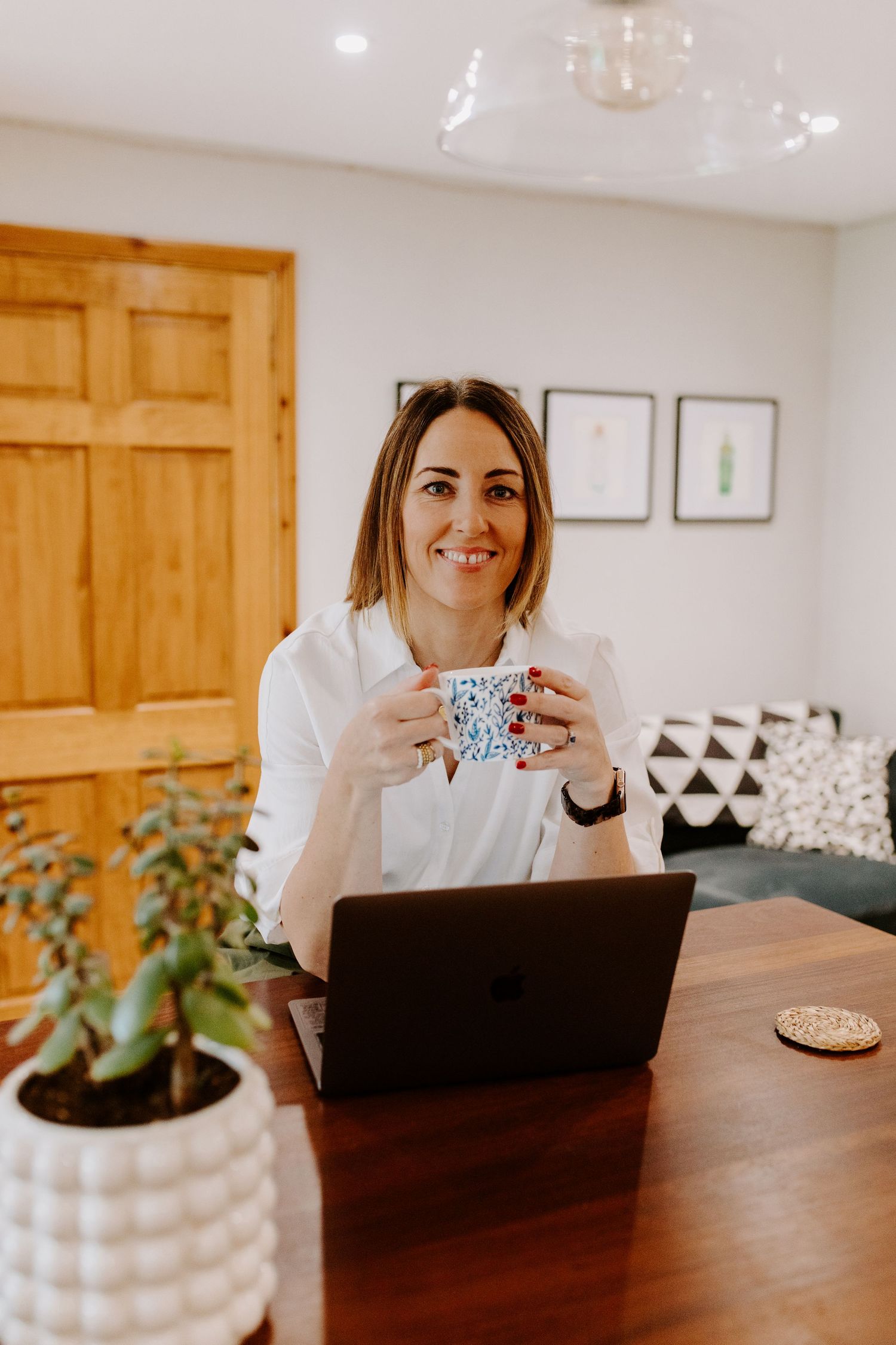 A person works at a wooden desk with a laptop and decorative plant in a bright modern home office setting.