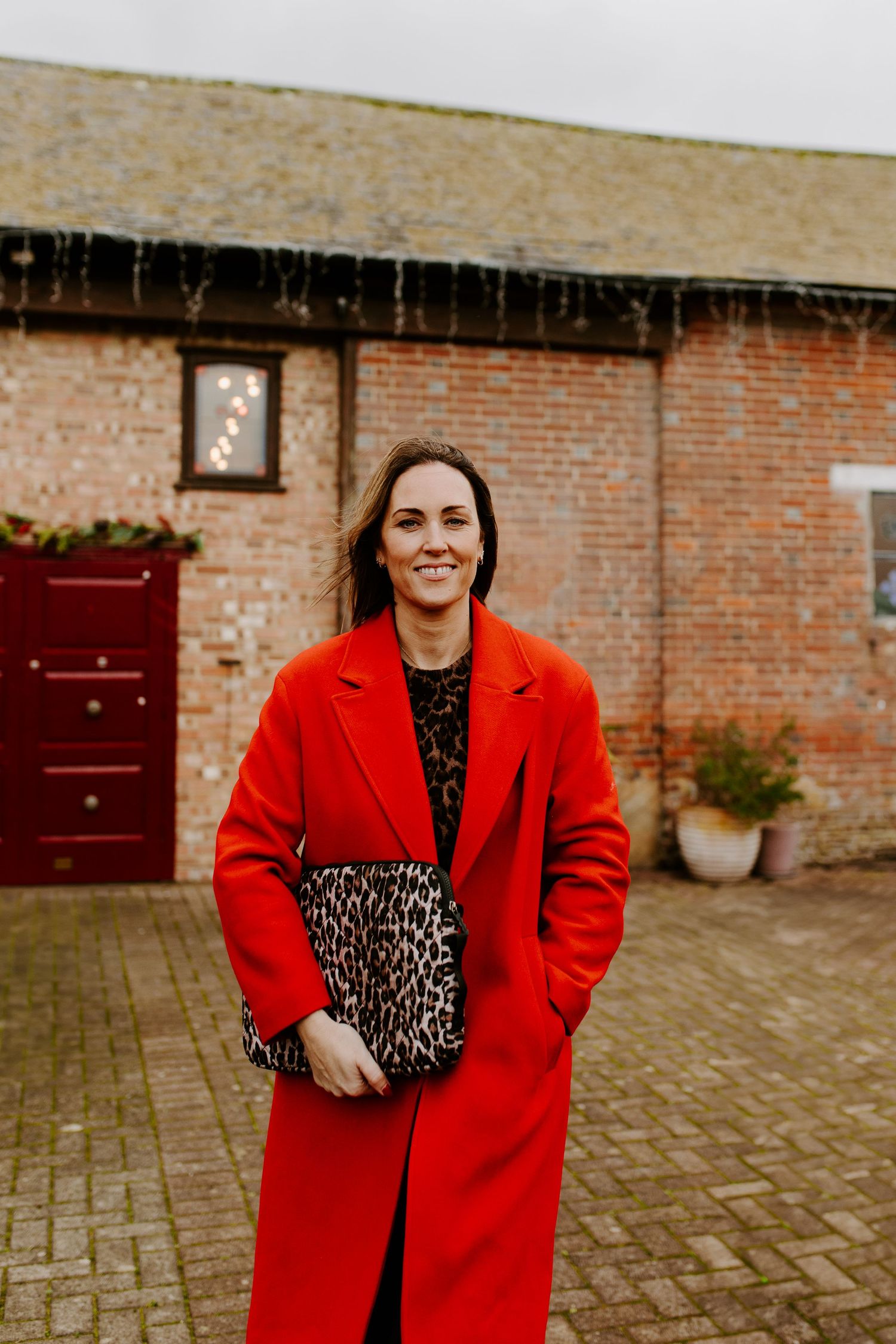 Person in bright red coat and polka dot dress posing against brick wall exterior.
