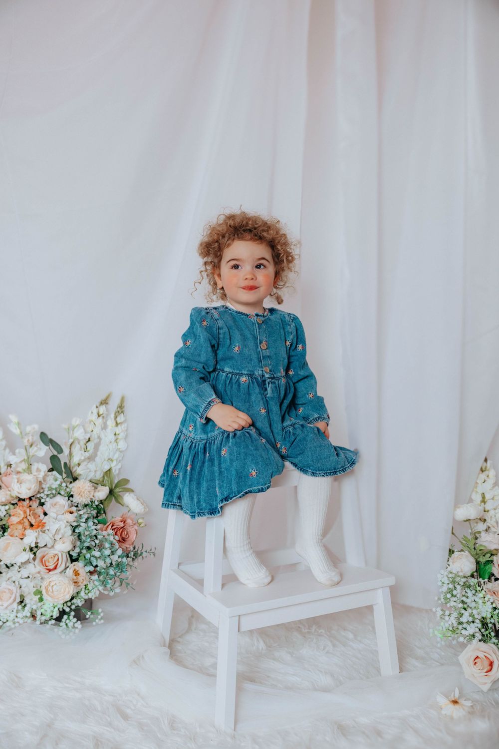 Child in blue denim dress standing on white step stool with floral decorations.