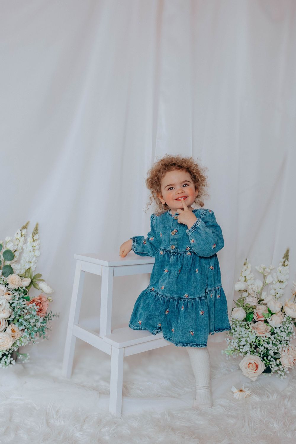 Playful toddler in blue dress posing with hand on chin next to white step stool.