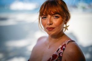 A close up portrait of someone with bangs wearing a red top against a soft blurred sky background.