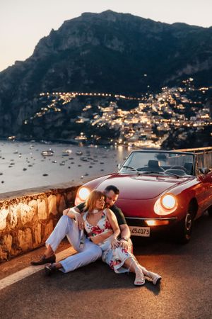 A couple sitting beside a vintage red convertible car overlooking the Amalfi Coast at sunset with twinkling city lights below.