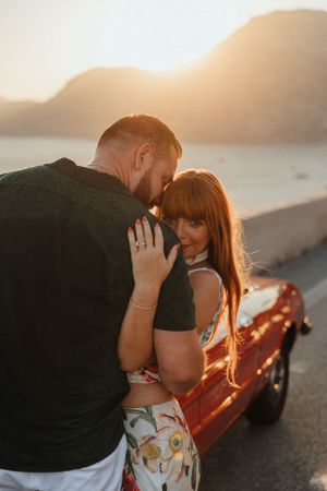 A romantic couple embraces next to a parked red car during golden hour sunset on a scenic mountain road.