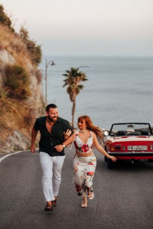 A couple runs joyfully down a coastal road at sunset with a red convertible parked behind them and a palm tree view.