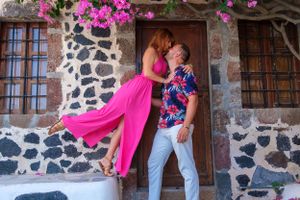 A romantic couple poses by a rustic stone wall, with a flowing pink dress creating dramatic movement in the scene.