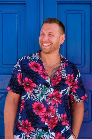 Someone wearing a colorful tropical print Hawaiian shirt with red hibiscus flowers stands smiling against a bright blue door.