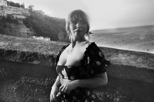 A person in a dress poses along a stone wall with a hillside and ocean visible in the black and white photograph.