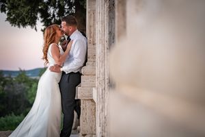 A romantic wedding couple embrace in a gentle pose by an elegant stone pillar at sunset.