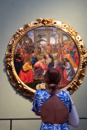 Museum visitor in blue backless dress views ornate gold-framed religious Renaissance painting on gallery wall.