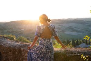 Person in floral dress standing at scenic overlook during golden sunset over rolling hills and countryside landscape.