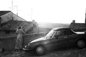Vintage black and white photograph of a convertible car parked on a cobblestone street at sunset with a figure standing nearby.