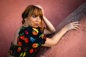 A person wearing a floral pattern dress with fruit designs leans against a pink textured wall in a stylized portrait shot.