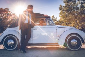 Classic white Volkswagen Beetle parked on a road with sunlight streaming through trees in golden-hour lighting.