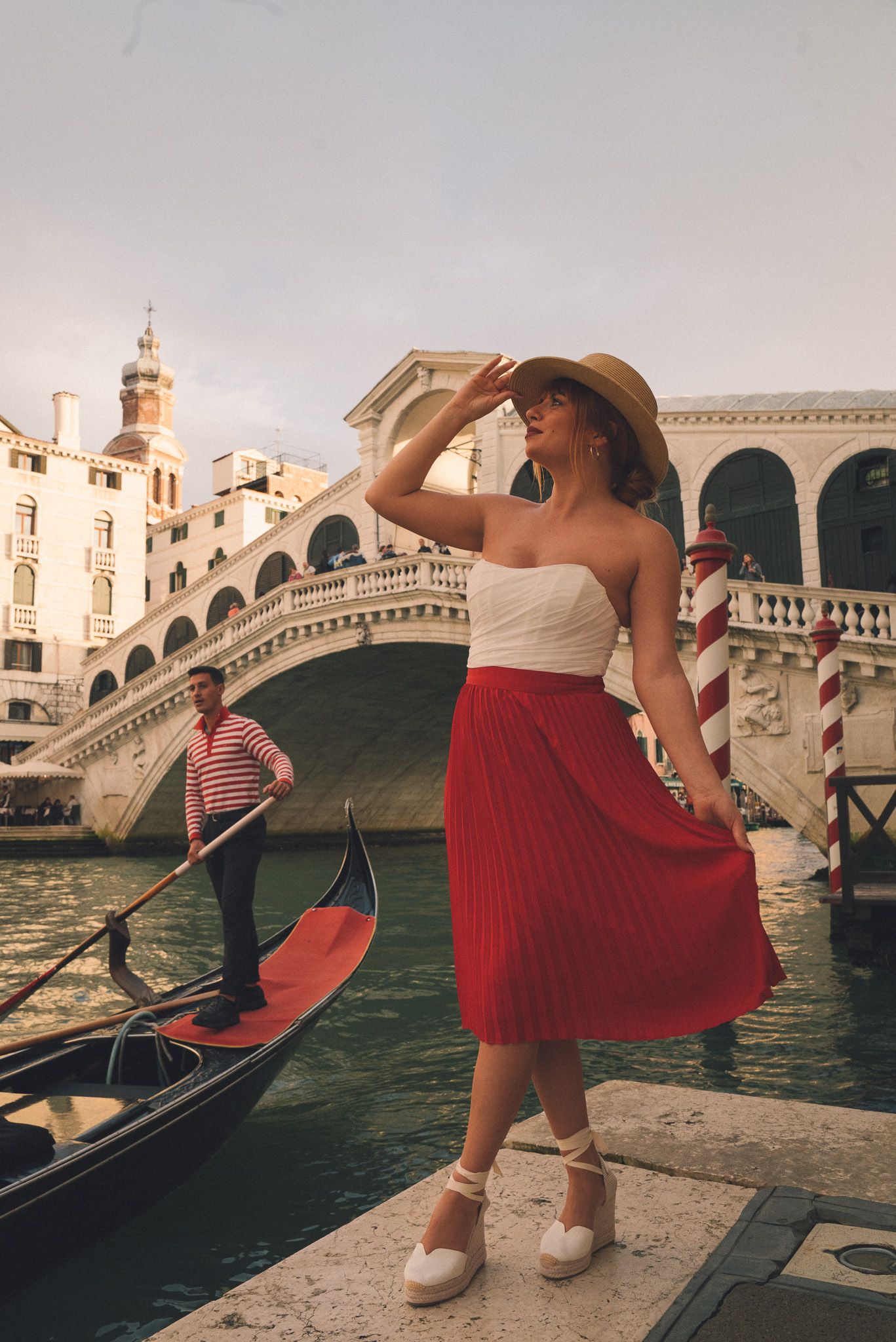 Tourist in red skirt and sun hat poses near Venice's iconic Rialto Bridge as gondola passes on Grand Canal.