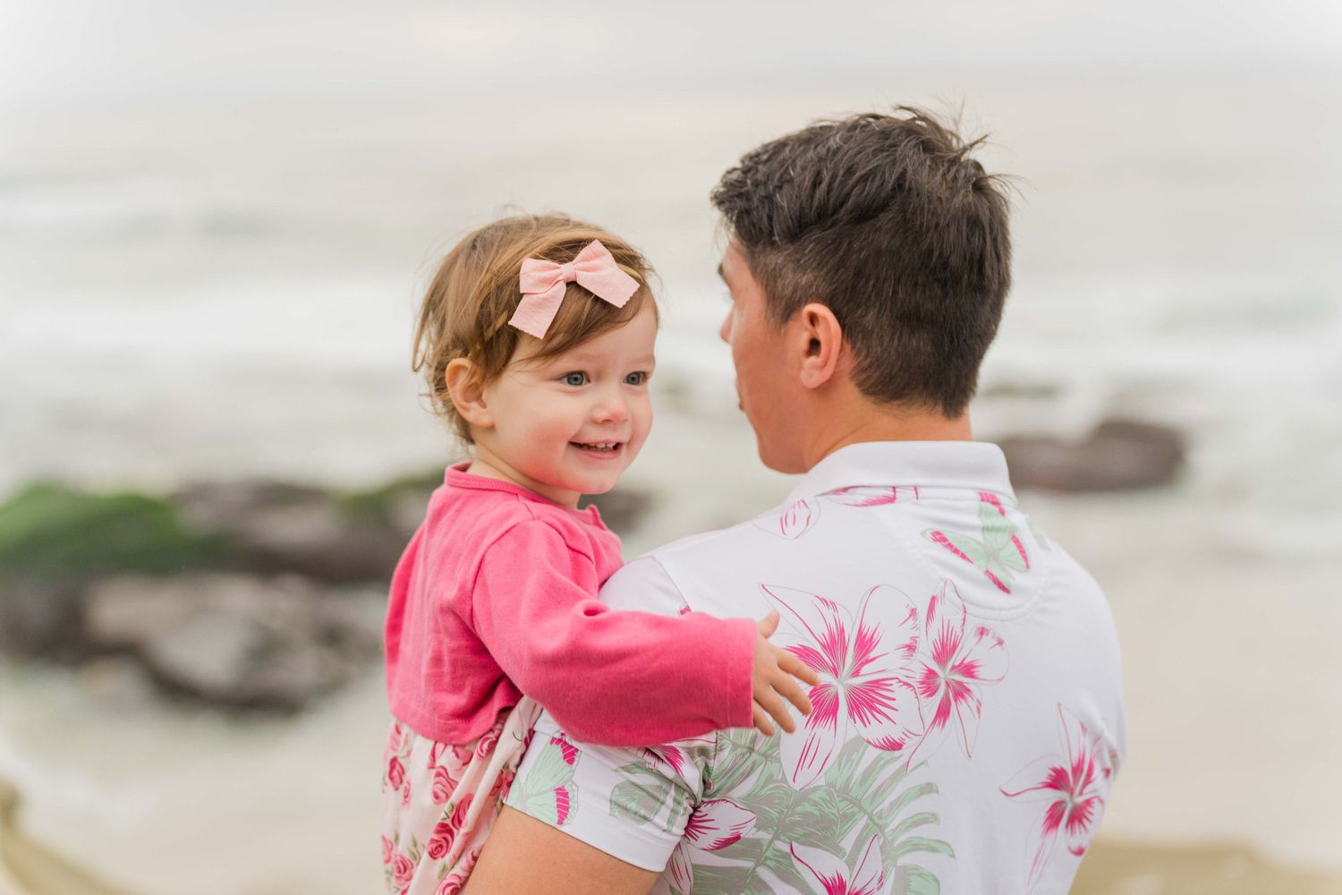 Parent and child sharing tender moment on rocky beach with ocean waves behind them.