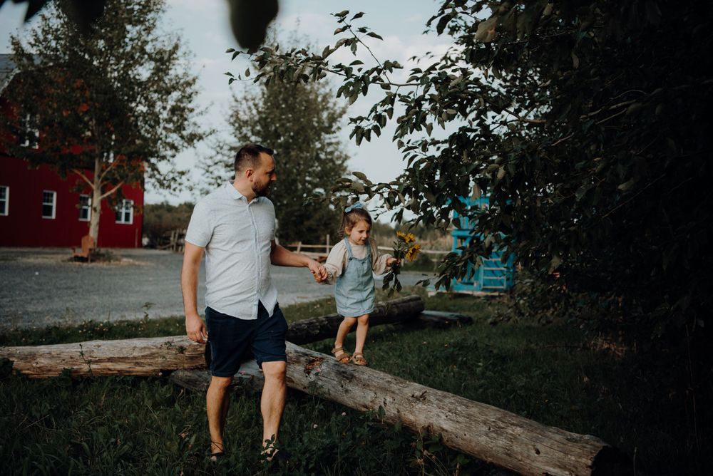 Parent and child balancing on log together near red barn.