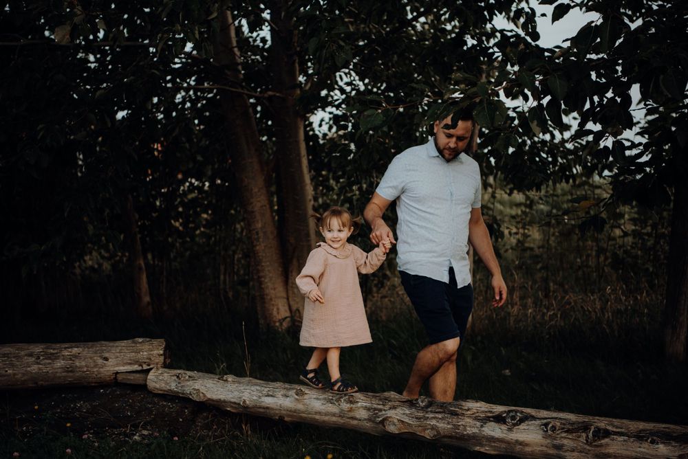 A parent and child walk together on a log in a wooded area at dusk.