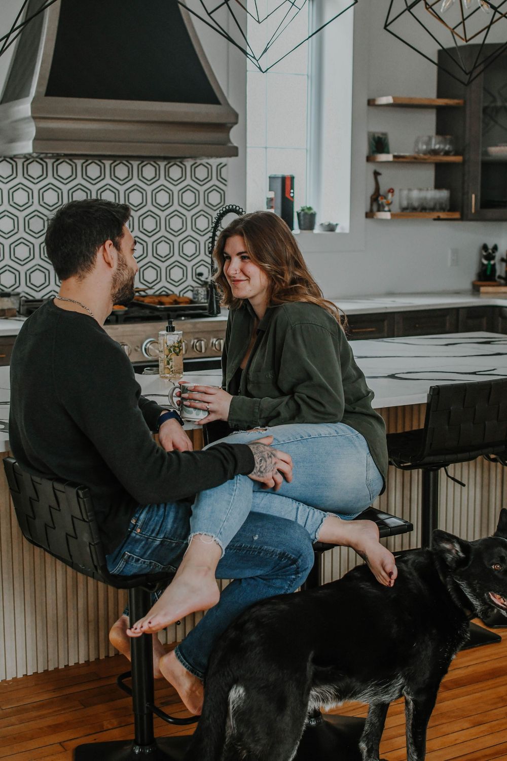 Couple sitting at kitchen counter with black dog, featuring geometric tile backsplash and modern farmhouse decor.