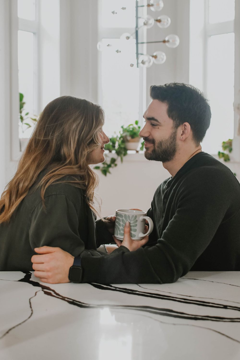 Couple in dark sweaters sharing an intimate moment while holding a mug in bright modern interior with hanging lights.