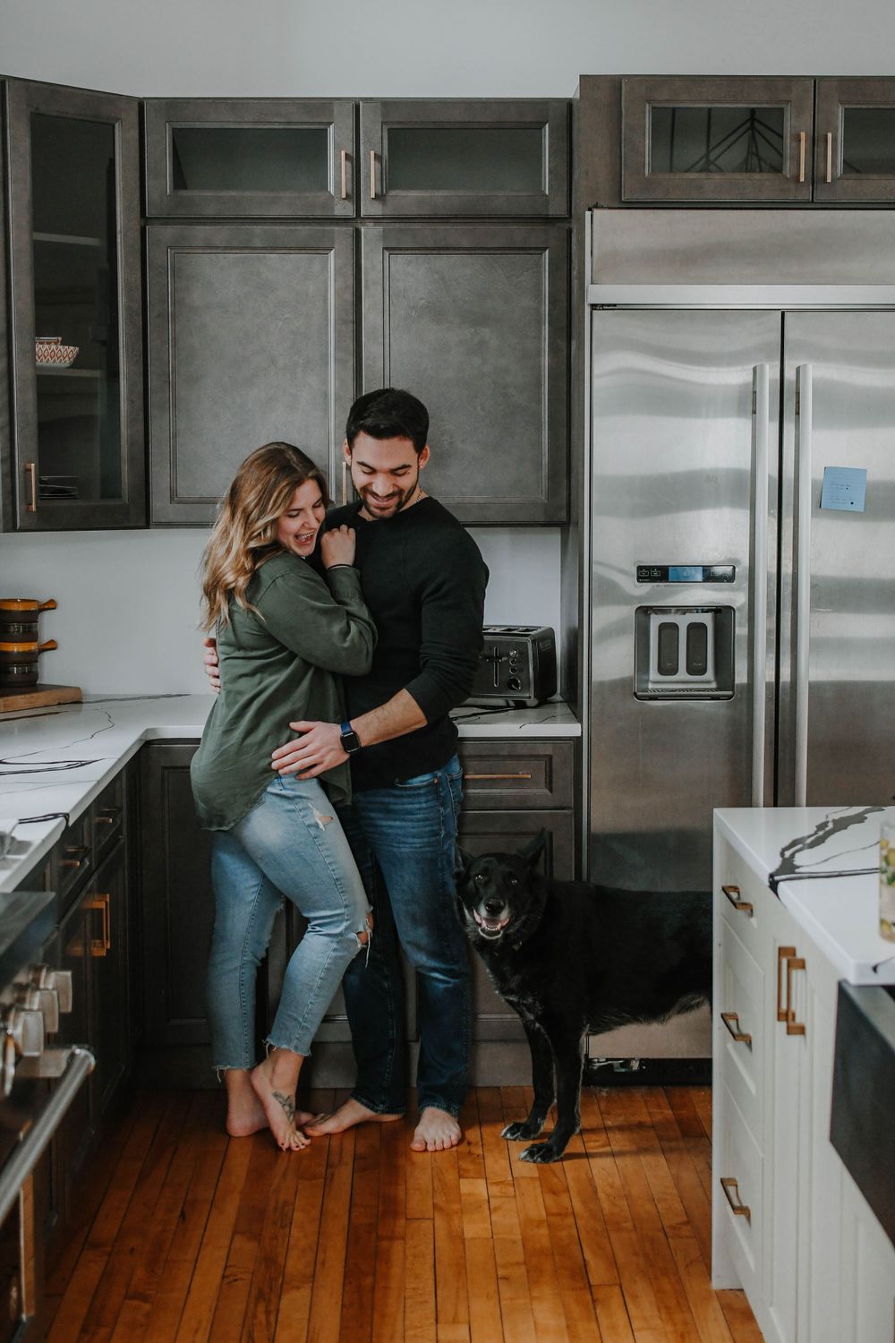 Couple embraces in modern kitchen with hardwood floors while black dog stands nearby.