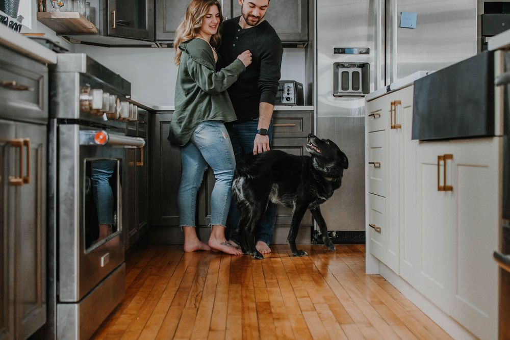 Couple standing barefoot in modern white kitchen with black dog on hardwood floor.
