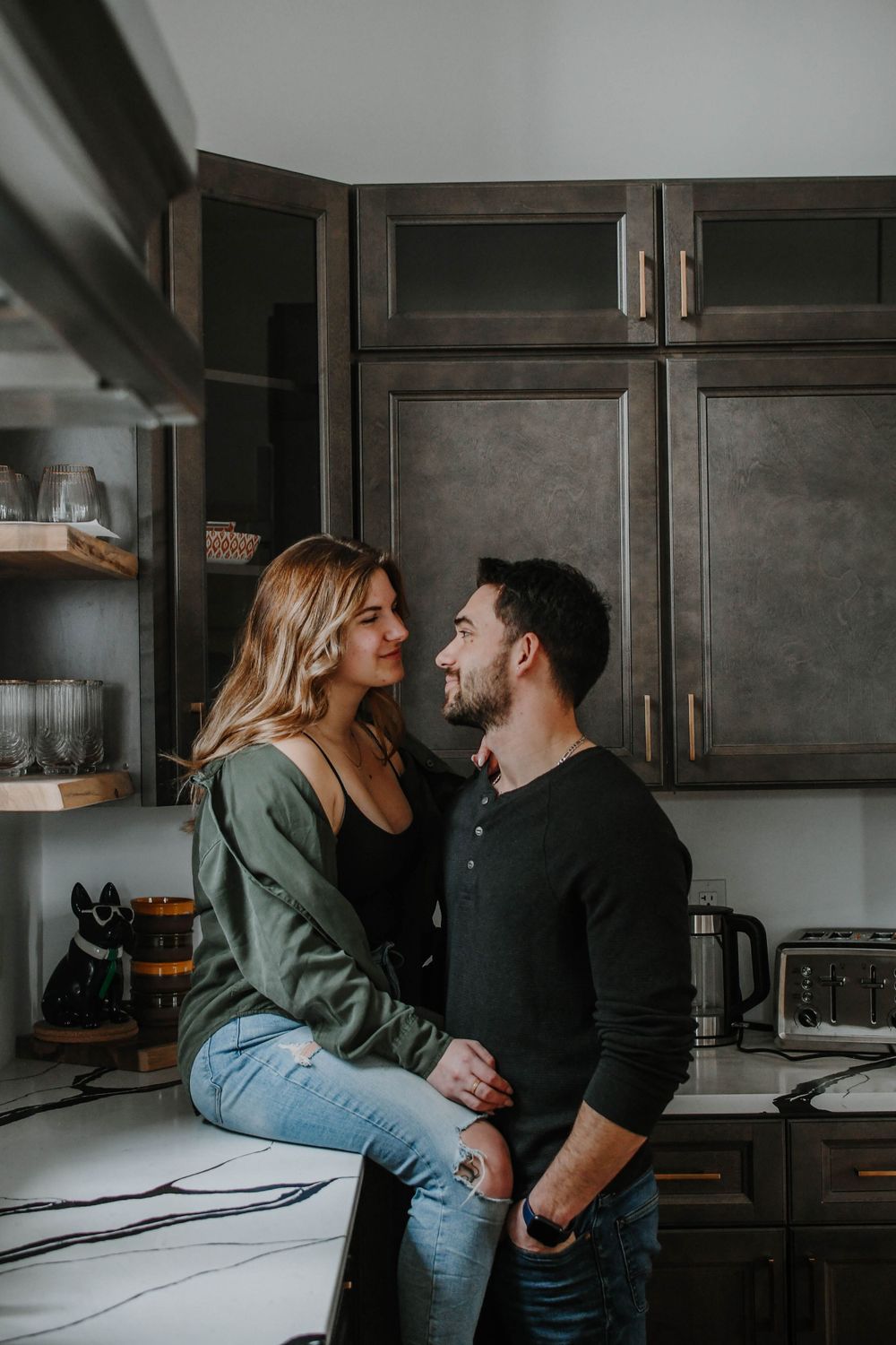 Couple sharing an intimate moment together in a moody kitchen with dark cabinets and natural lighting.