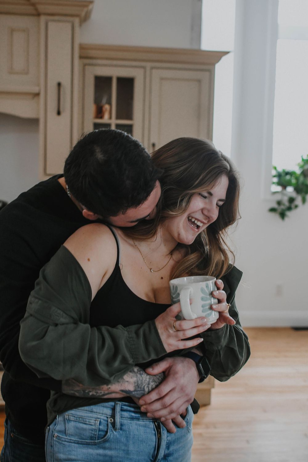 Couple sharing an intimate moment in their kitchen as person holds coffee mug while being embraced from behind.