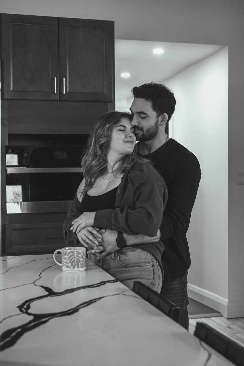 Romantic black and white photo of a couple embracing in their kitchen with a coffee mug on the counter.