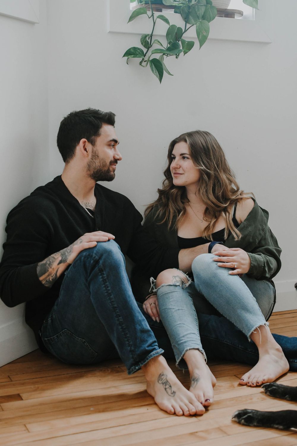 Intimate couple sits together on wooden floor with a green plant hanging above them, sharing a casual moment.