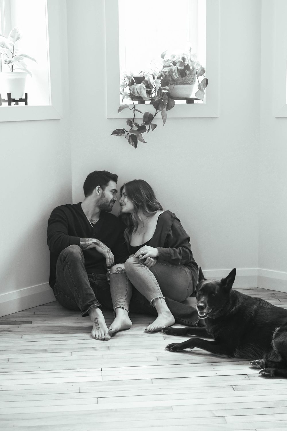 A couple sits intimately on wooden floor with black dog beside them and hanging plants in white minimalist room.