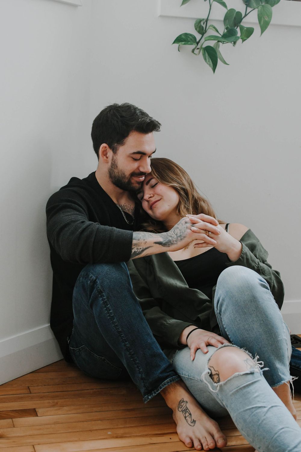 Couple sharing an intimate moment while sitting on wooden floor against white wall with green plant decoration.