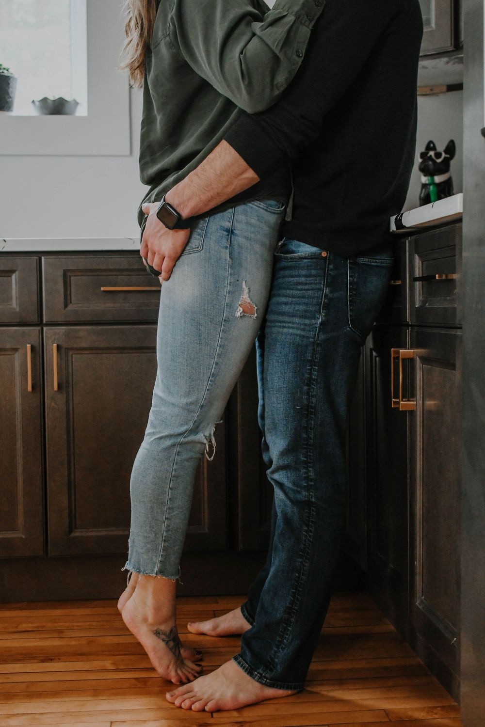 Couple in casual attire standing barefoot together in kitchen with dark cabinets and hardwood floors.