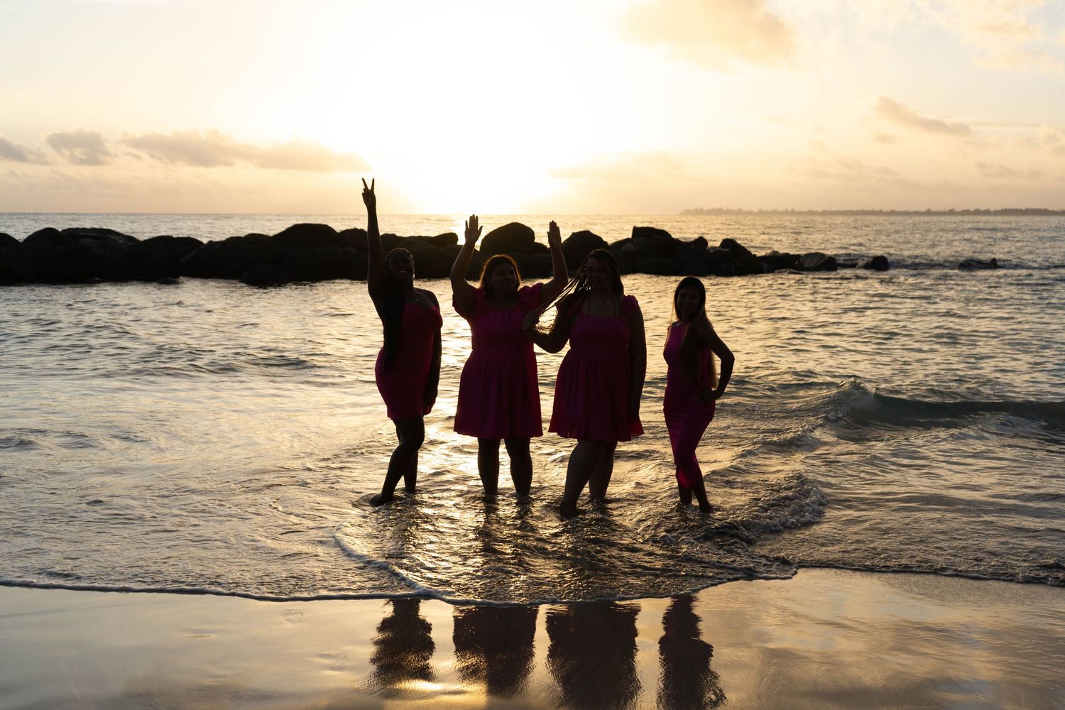 Silhouettes of four women in matching dresses celebrating on a beach at golden sunset.